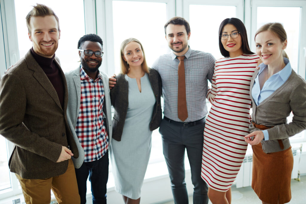 Three pretty businesswomen and three bearded businessmen standing against panoramic office window and looking at camera with wide smiles