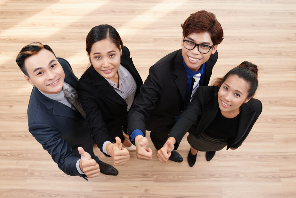 Directly above view of cheerful Asian white collar workers looking at camera with wide smiles and showing thumbs up