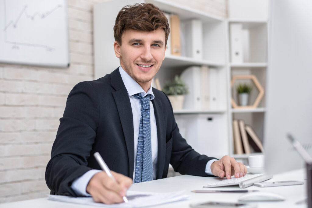 Portrait of handsome young businessman looking at camera smiling cheerfully while working at desk in modern office