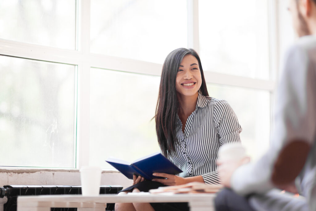 Pretty Asian manager looking at her male colleague with wide smile while having business meeting in office lobby with panoramic windows, portrait shot